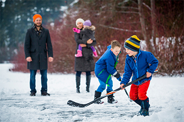 Two kid brothers playing ice hockey on a lake while their parents watch. 